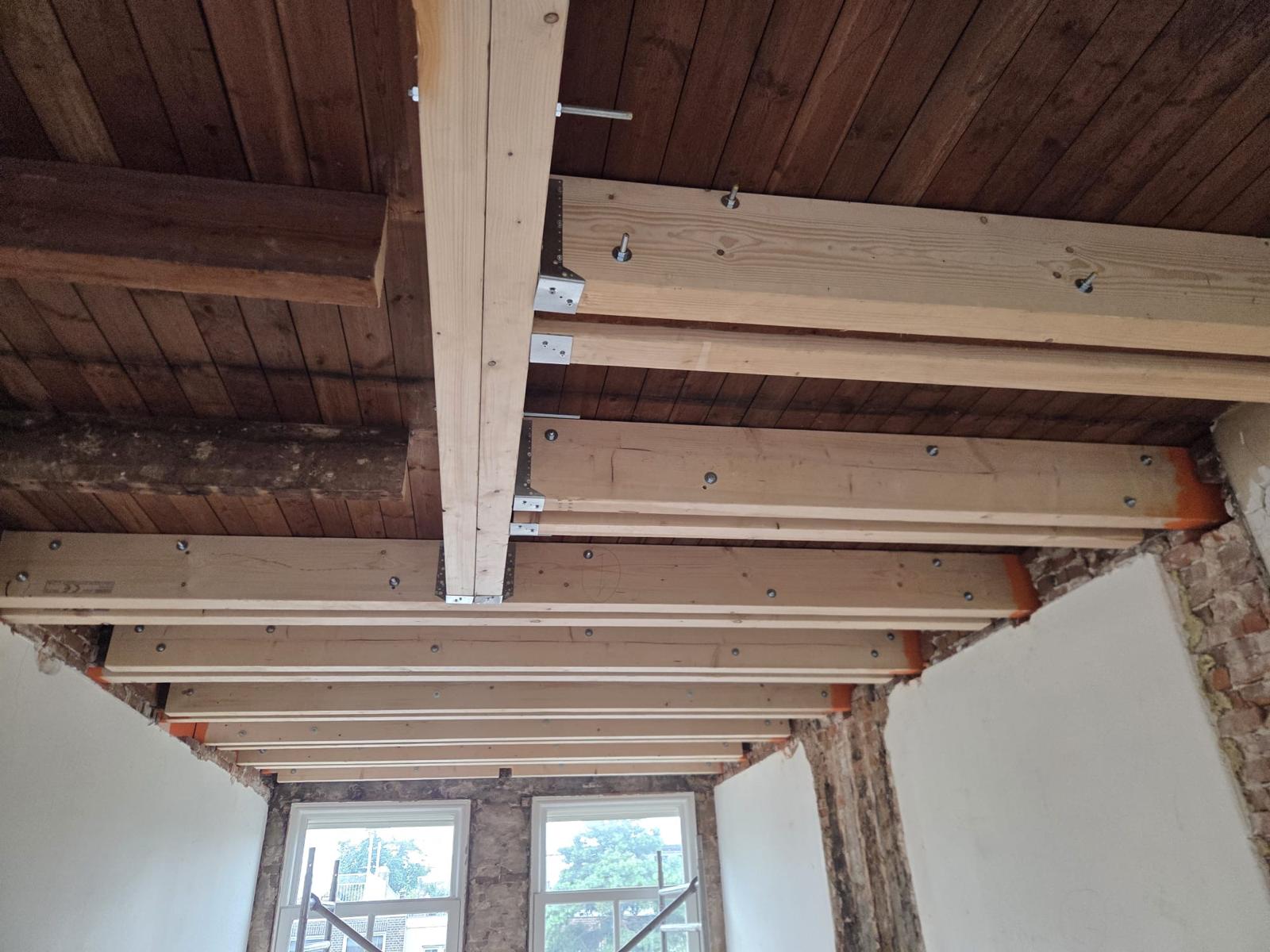 Bestaande houten balklaag versterkt met nieuwe balken en stalen balkschoenen, onder een oud houten plafond