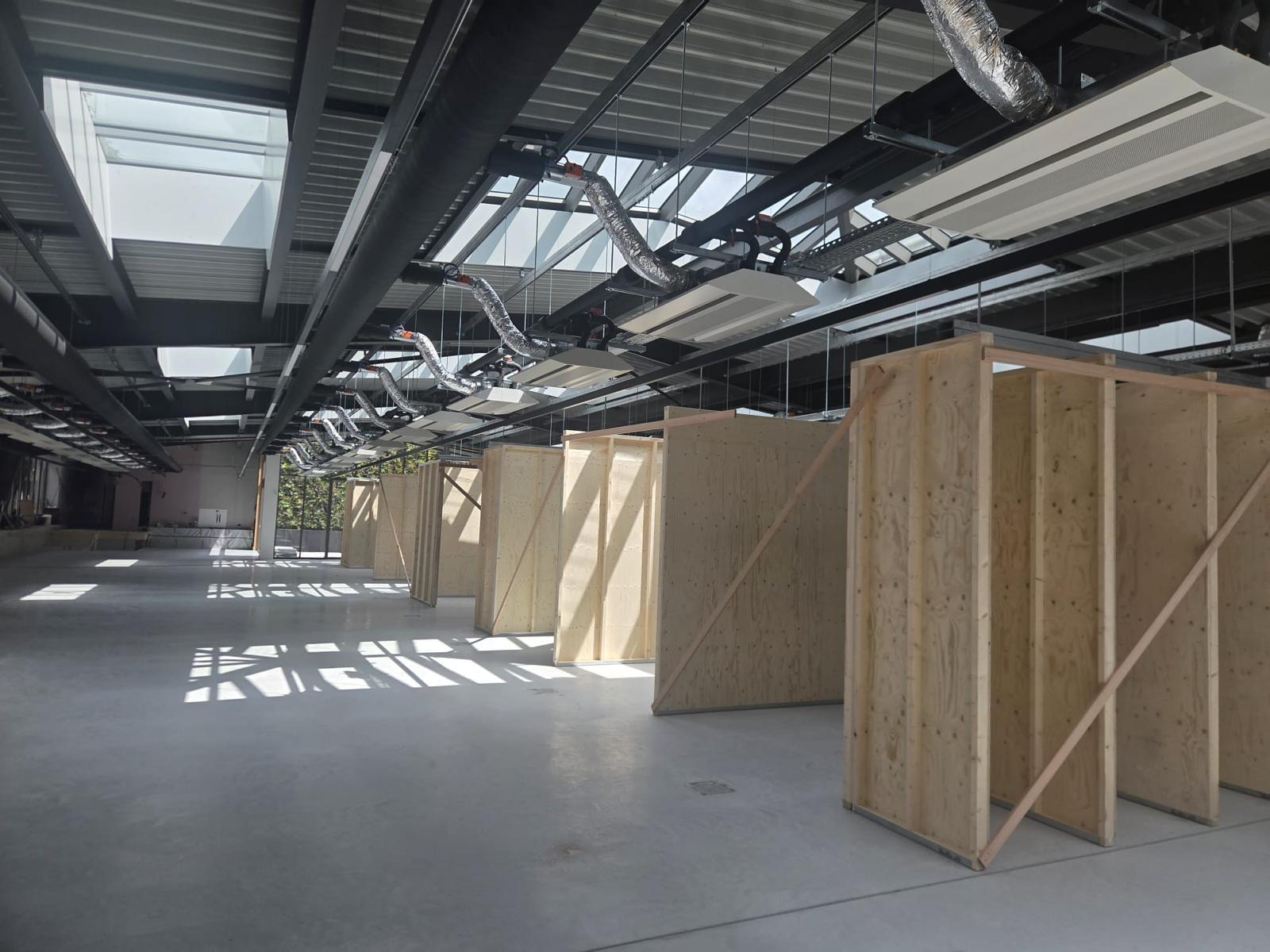 Rij plywood cabines in een lichte bedrijfshal met zichtbare ventilatiekanalen en verlichting
