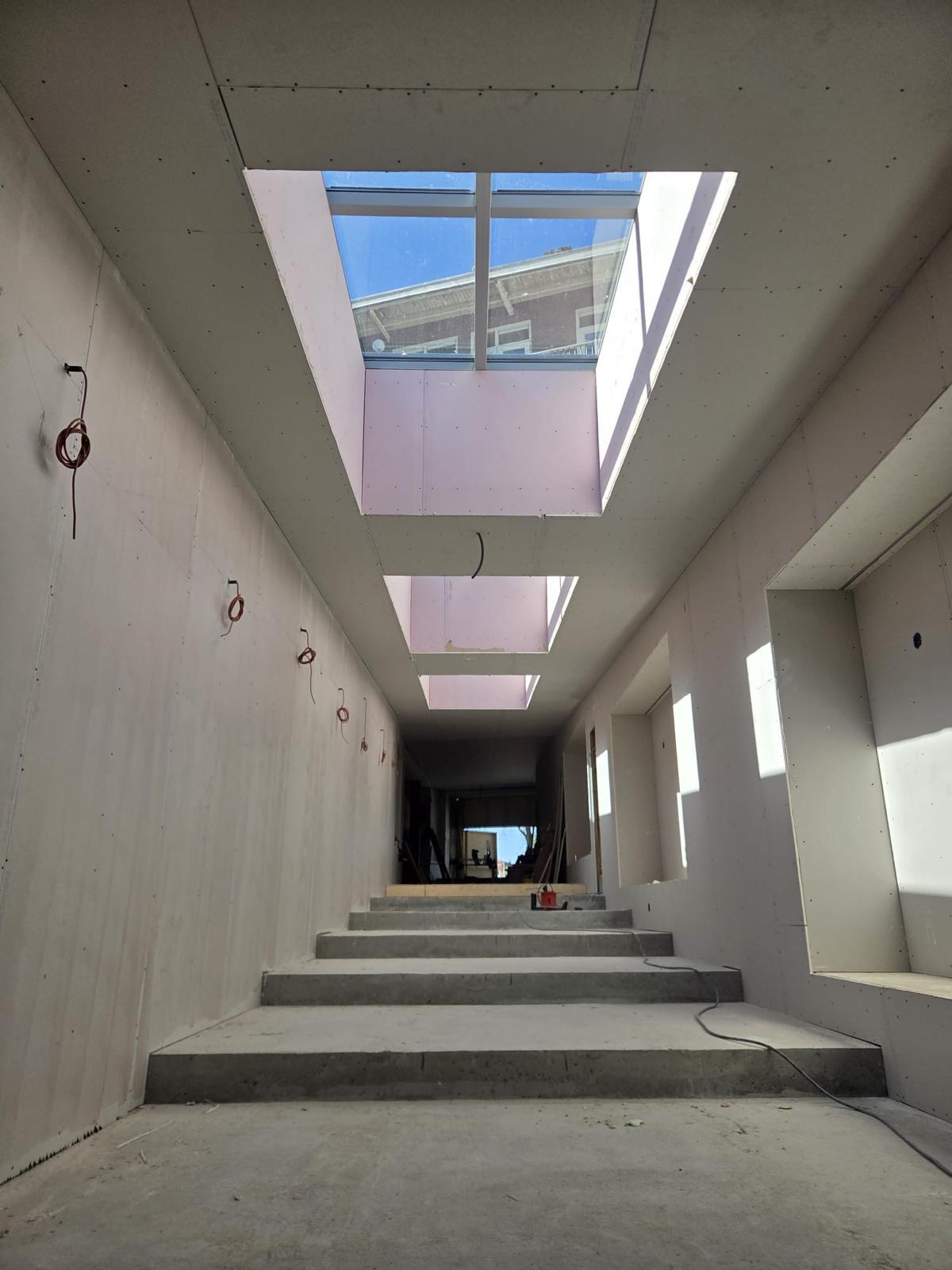 Gang met daglichtstraat in het plafond, strak afgewerkte gipswanden en een betonnen trap