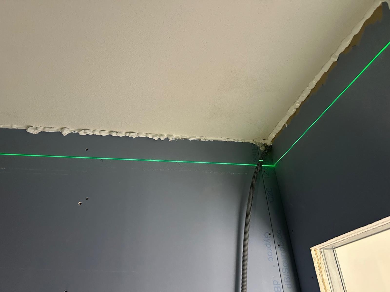 Groene laserlijn van een kruislijnlaser langs de plafondaansluiting van een grijze Gyproc dB SoundBloc wand